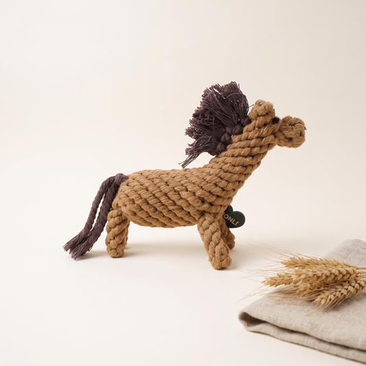 A rope horse dog toy made from natural tan rope with a dark brown mane and tail, standing on a light beige surface next to folded linen and wheat stalks.
