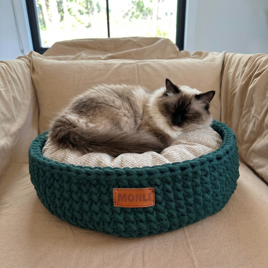 Cat lying on a green knitted pet bed with 'MONLI' branding on a beige couch.