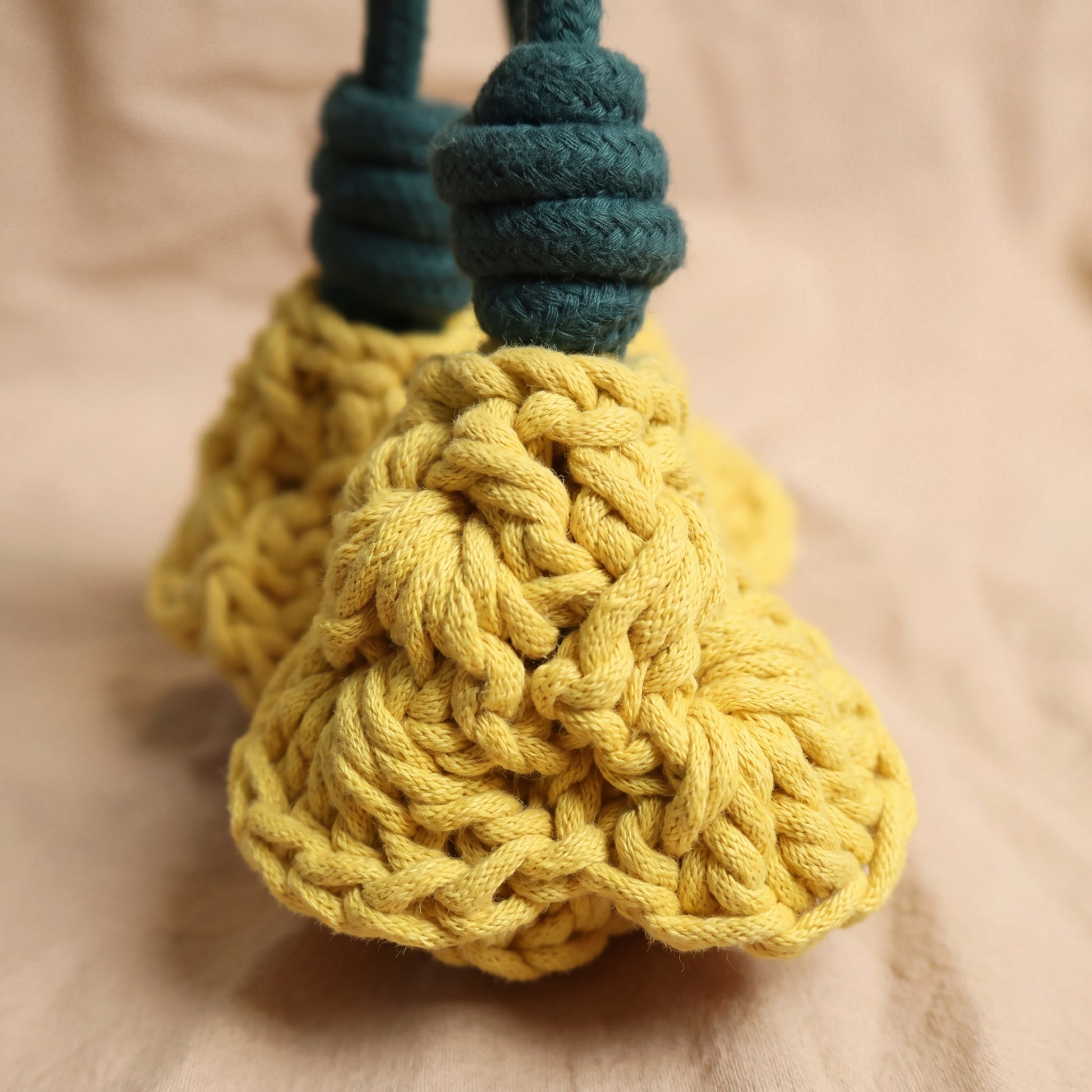 A close-up of a yellow knitted flower with a dark teal rope handle, resting on a soft beige fabric background.