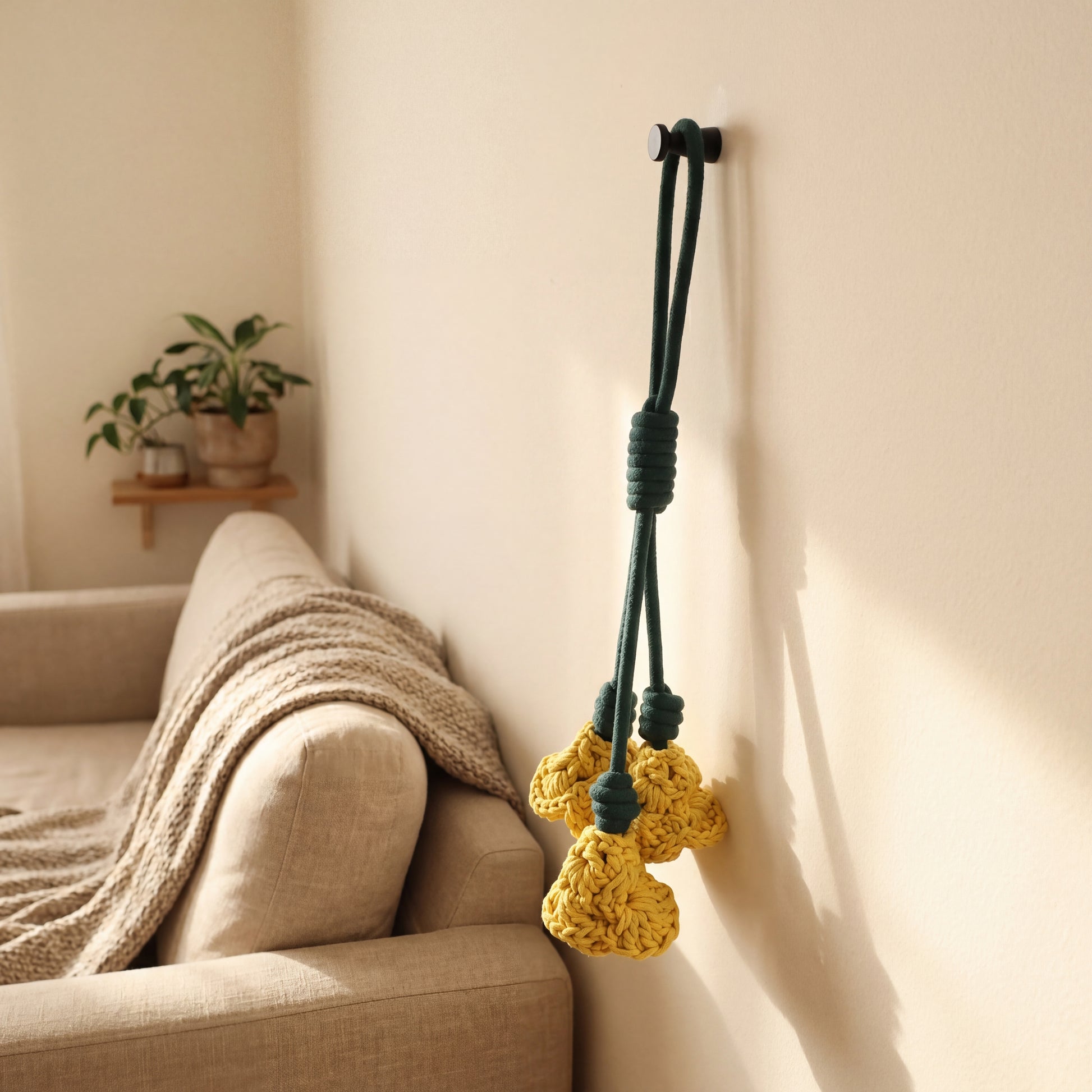 A dark green wall hook with three knotted yellow flowers hanging on a beige wall next to a sofa with a throw blanket.