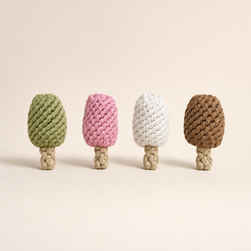 Four knotted ice cream bar rope dog toys shaped like pinecones in sage green, pink, off-white, and brown, arranged in a row on a light beige background.
