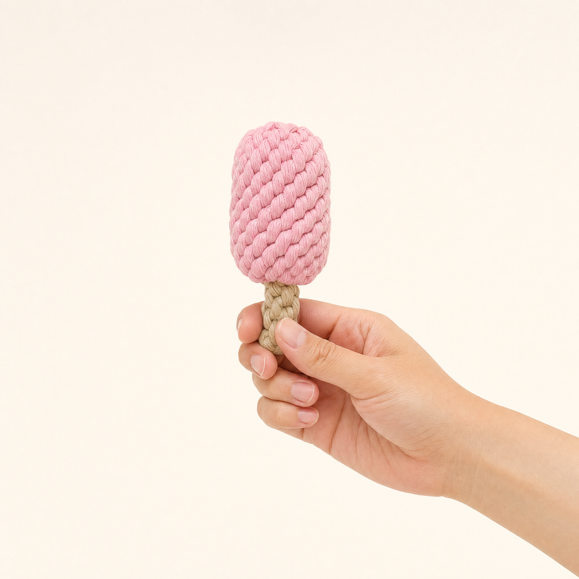 A hand holding a pink textured rope toy shaped like a popsicle with a beige rope stick, set against a solid light cream background.