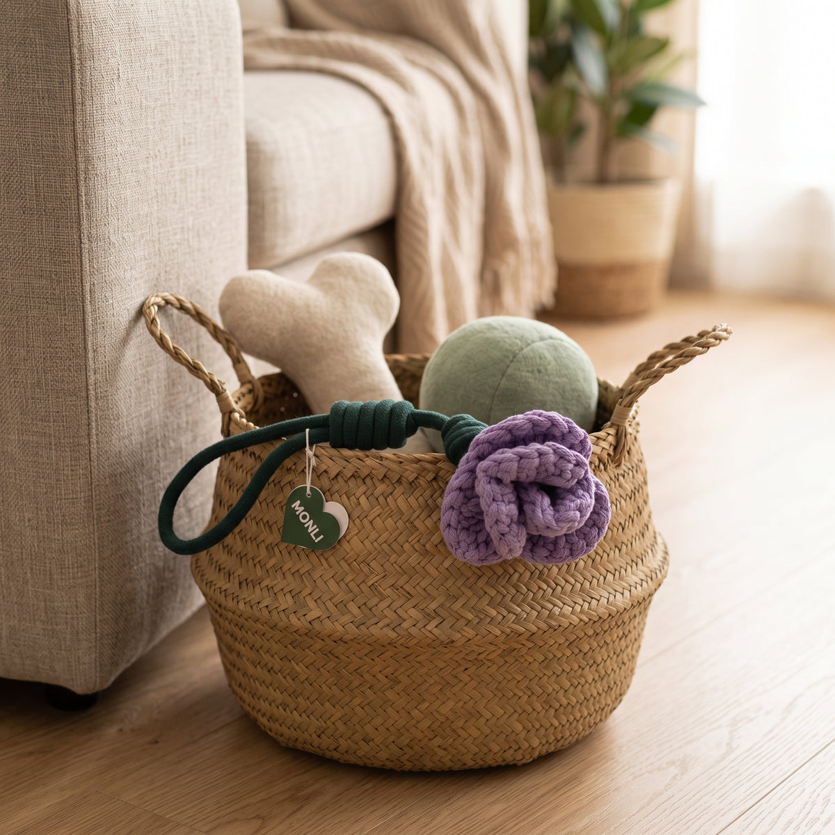A woven natural-fiber storage basket on a wooden floor beside a sofa, holding dog toys including a cream bone-shaped toy, a green ball, a dark green rope toy, and a purple knitted flower toy.